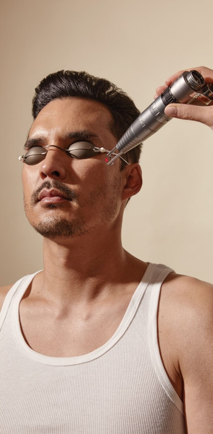 A man wearing protective eye shields and a white tank top, receiving a facial laser treatment near his temple from a handheld device, with a red laser light visible.