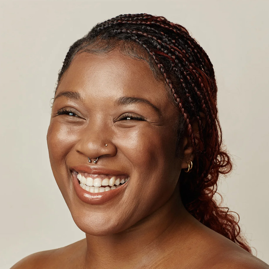 A joyful woman with braided hair and glowing skin, smiling widely at the camera, showcasing healthy, radiant complexion.