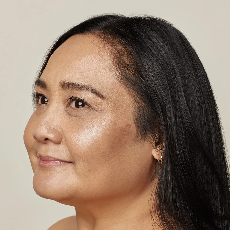 A side-profile portrait of a woman with long dark hair and healthy, glowing skin, looking to the left with a subtle smile, highlighting her natural complexion.