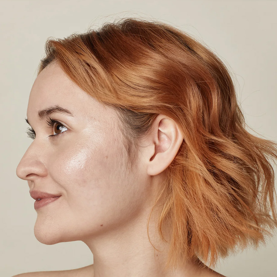 A side-profile portrait of a person with short, wavy reddish-blonde hair and glowing, hydrated skin, looking upwards with a subtle smile.