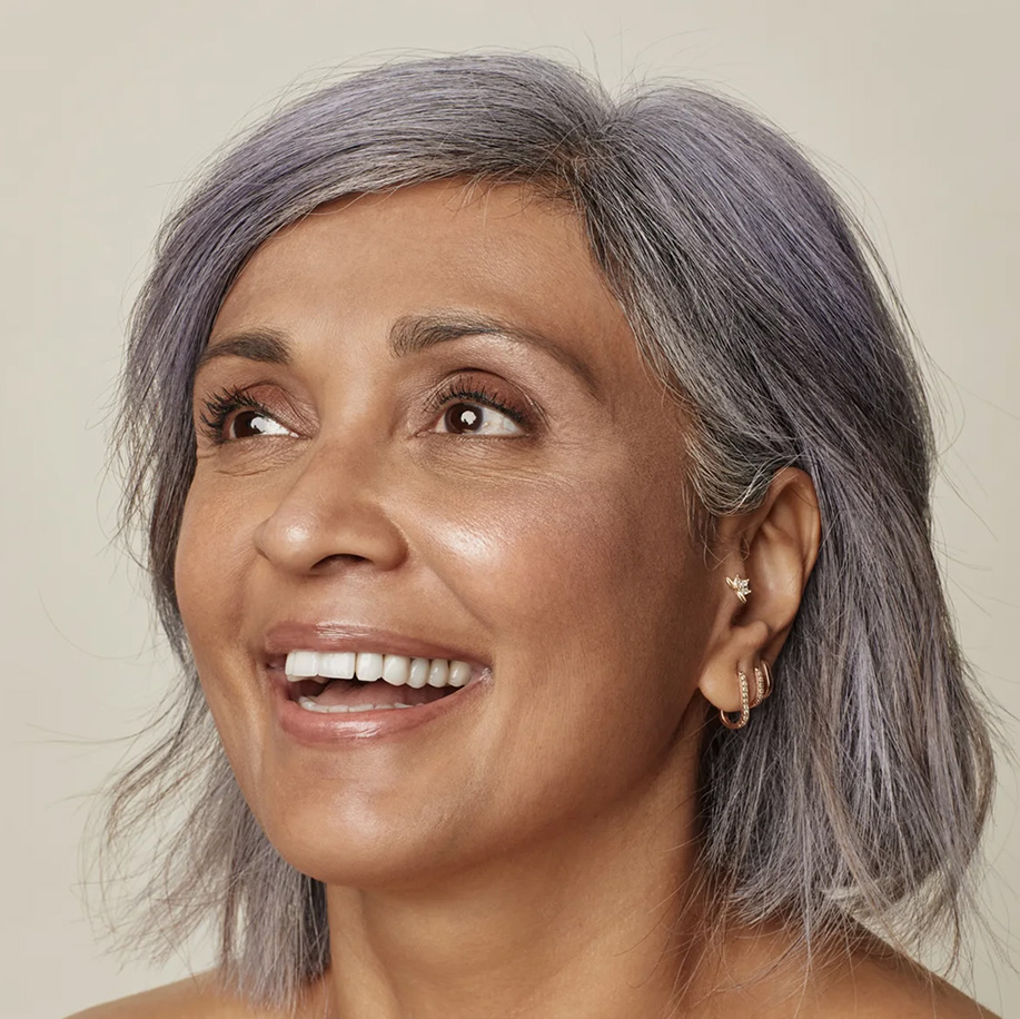 A smiling woman with short, stylish grey hair and clear, glowing skin, wearing delicate earrings, looking up and to the right with a joyful expression.