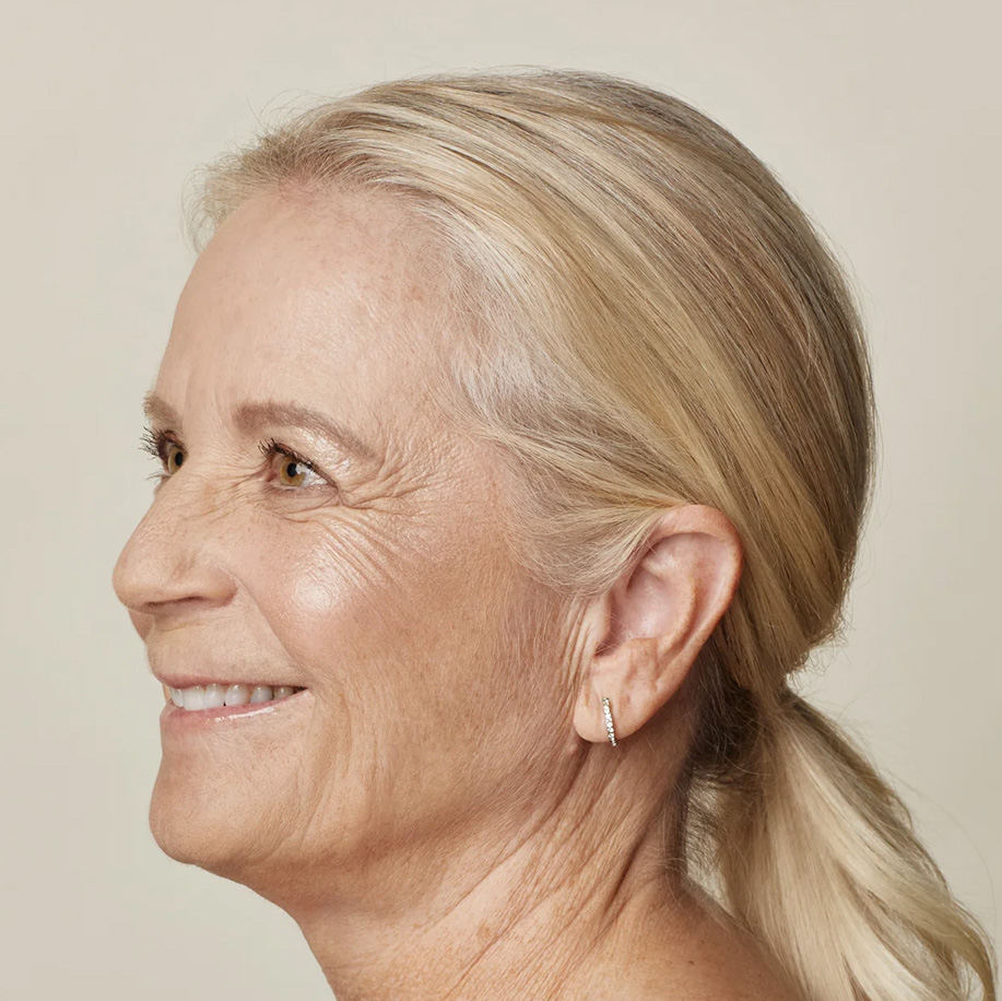 A side-profile portrait of a smiling older woman with blonde hair in a low ponytail and clear, glowing skin, wearing a small hoop earring.